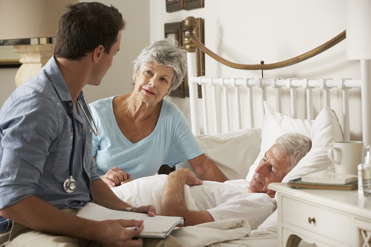 Doctor On Home Visit Discussing Health Of Senior Male Patient With Wife In Bed.