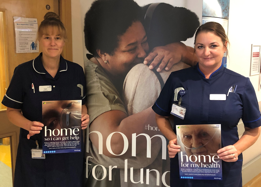 Two nurses stand in front of a banner with the words 'home for lunch', and hold campaign posters up outside a hospital ward.