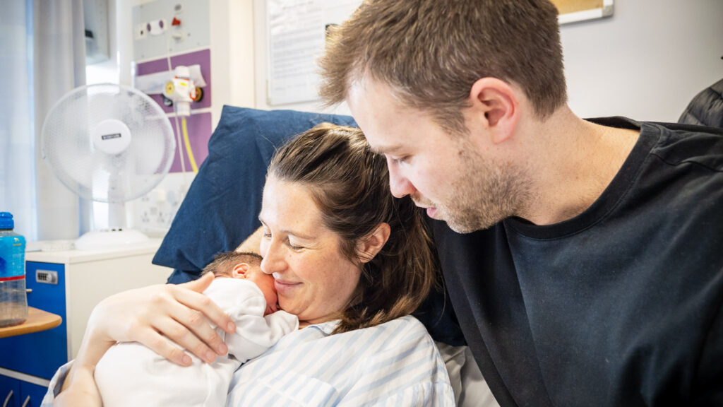 A mother holding a newborn baby in a hospital bed with her partner by her side