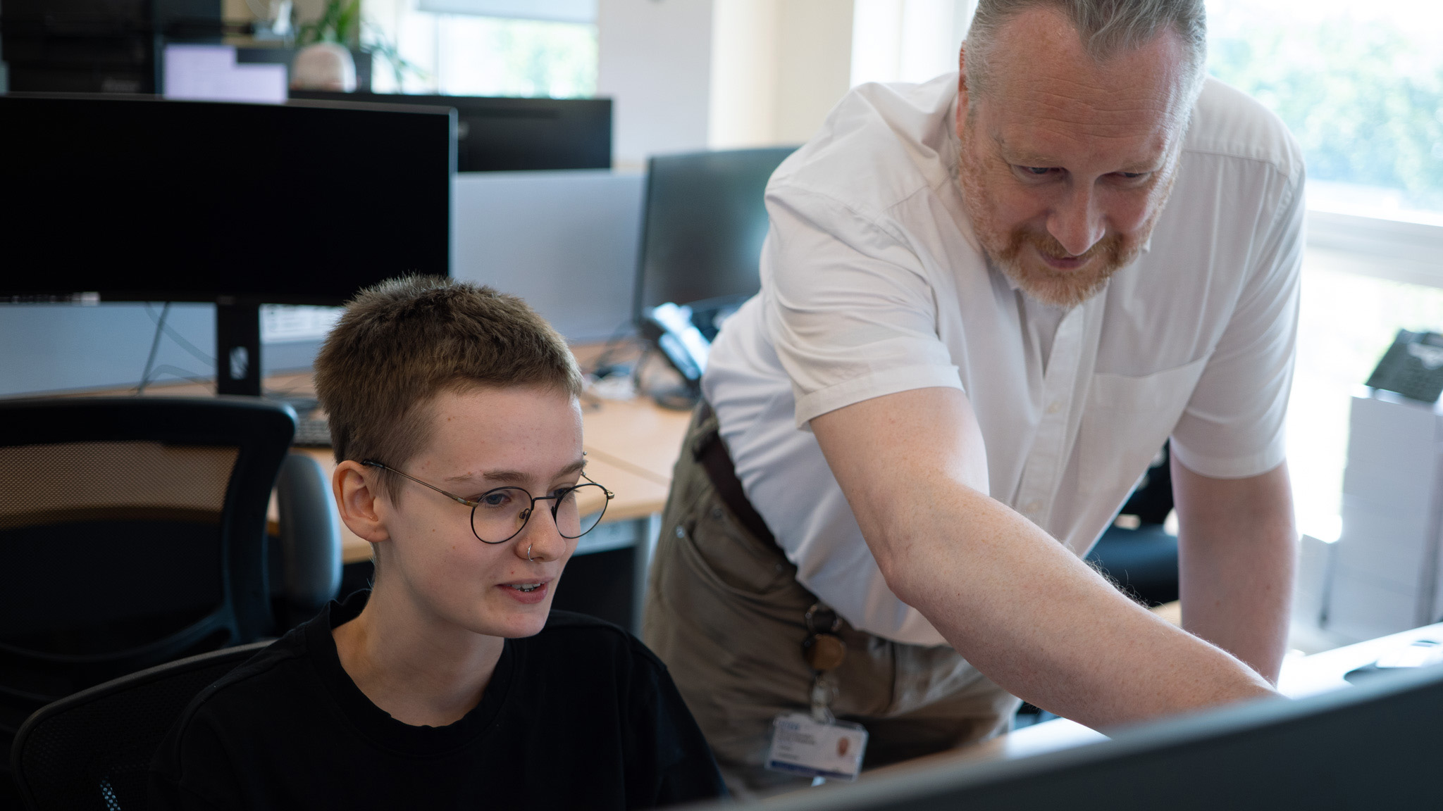 Photograph of young apprentice looking at computer screen with work colleague standing beside her pointing at the screen. Background shows lots of computer screens, tables and chairs.