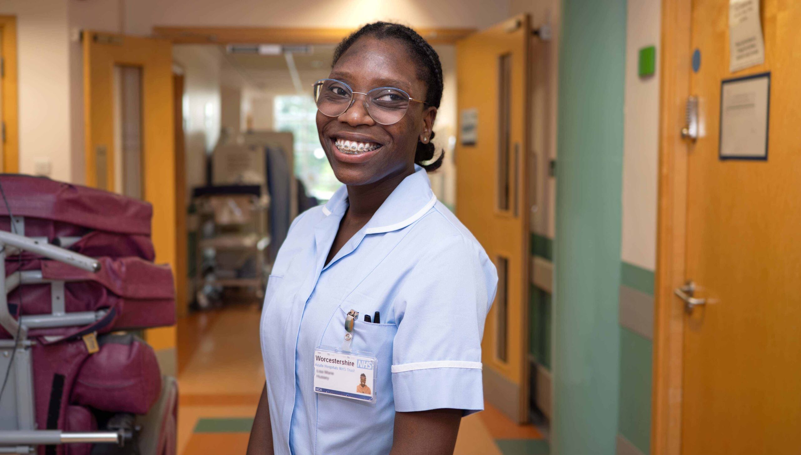 Photograph of a Support Worker smiling at work. Standing in the corridor of hospital. Door to ward open behind her.