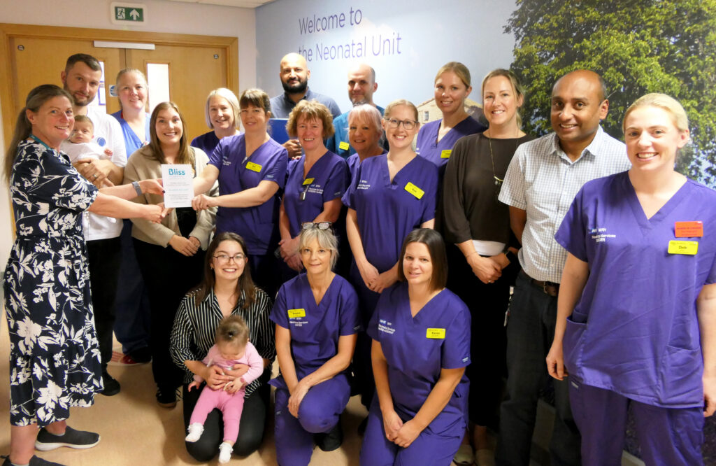 A large group of neonatal staff and families gather outside the Neonatal Unit, with two people holding a Bliss accreditation certificate. Two babies are pictured with their parents.