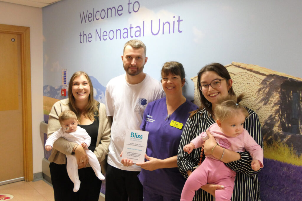 A group stands outside the Neonatal Unit, with two holding babies and a neonatal staff member holding a Bliss Baby Charter Gold accreditation certificate.