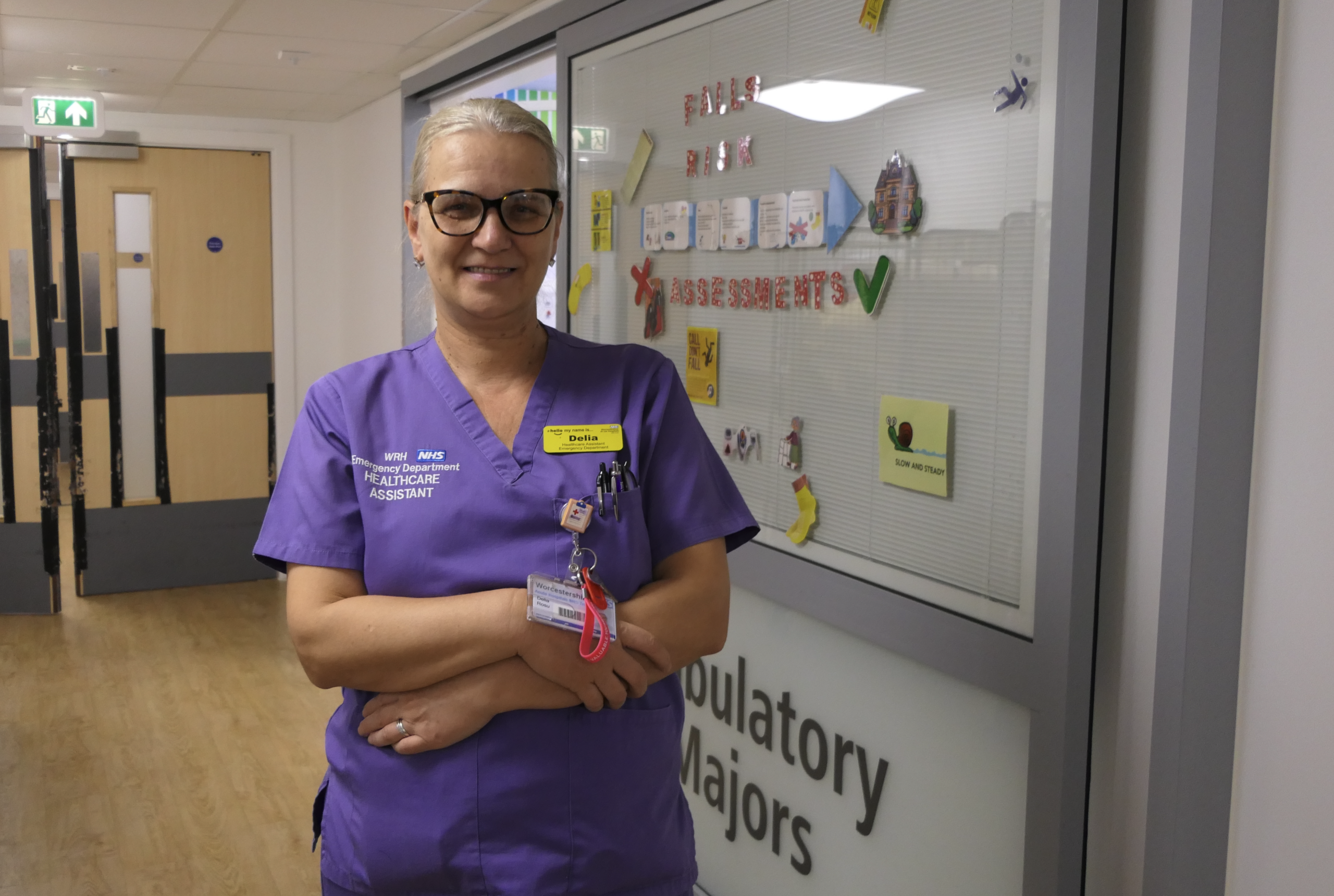 A hospital healthcare assistant in purple scrubs stands with arms folded in a corridor beside a glass display promoting falls risk assessments.