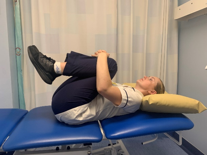 A picture of a person lying on their back on a bed, bringing both legs up and hug it towards the chest.