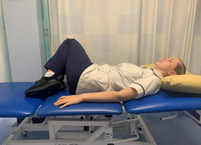 A picture of a person lying on their back on a bed, bending their knees to enable to plant both feet on the bed. Placed hands by their side, palms facing down. Slowly, roll knees from side to side.