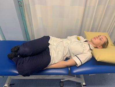 A picture of a person lying on their back on a bed, bending their knees to enable to plant both feet on the bed. Placed hands by their side, palms facing down. Slowly, roll knees from side to side.