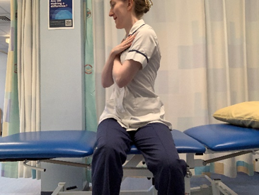 A picture of a person sitting on the side of the bed. Crossed their hands over the chest. Slowly, turn upper body round to the left and right.