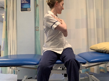 A picture of a person sitting on the side of the bed. Crossed their hands over the chest. Slowly, turn upper body round to the left and right.