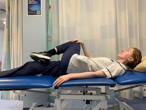 A picture of a person lying on their back on a bed, bend one knee and with the hand on the opposite side, gently pull the knee over towards the other leg.
