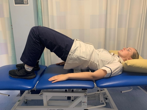 A picture of a person lying on their back on a bed with their knees bent. Slowly lift the hips away from the bed toward the ceiling